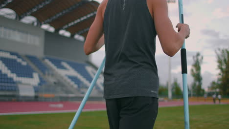 Ein-Speerwerfer-Läuft-Mit-Speeren-Durch-Das-Stadion-Und-Sammelt-Sie-Ein.-Gehen-Sie-Mit-Einem-Speer-In-Der-Hand-Vor-Dem-Hintergrund-Der-Stadiontribüne.-Das-Training-Des-Olympiasiegers.